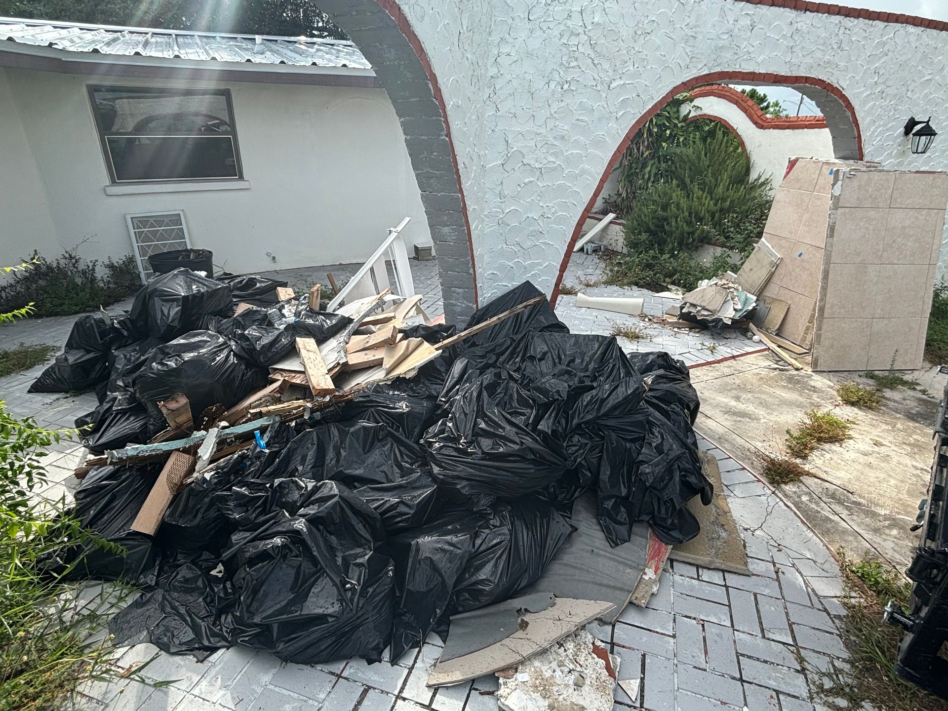 Pile of black trash bags, wood debris, and construction materials near a white stucco building with an arched entrance.
