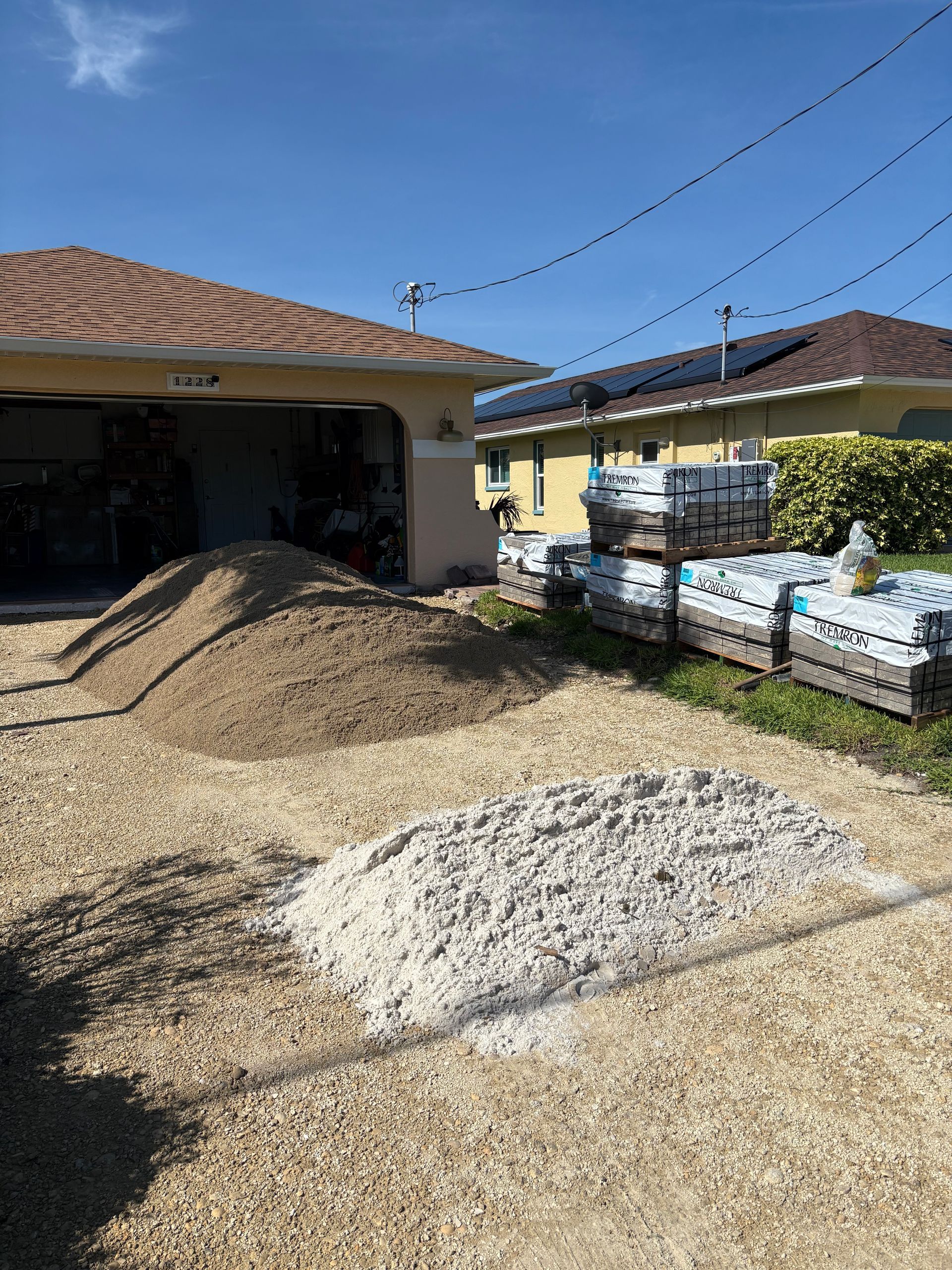 Piles of gravel and sand in a driveway, with stacked pavers in the background, near a house with solar panels.