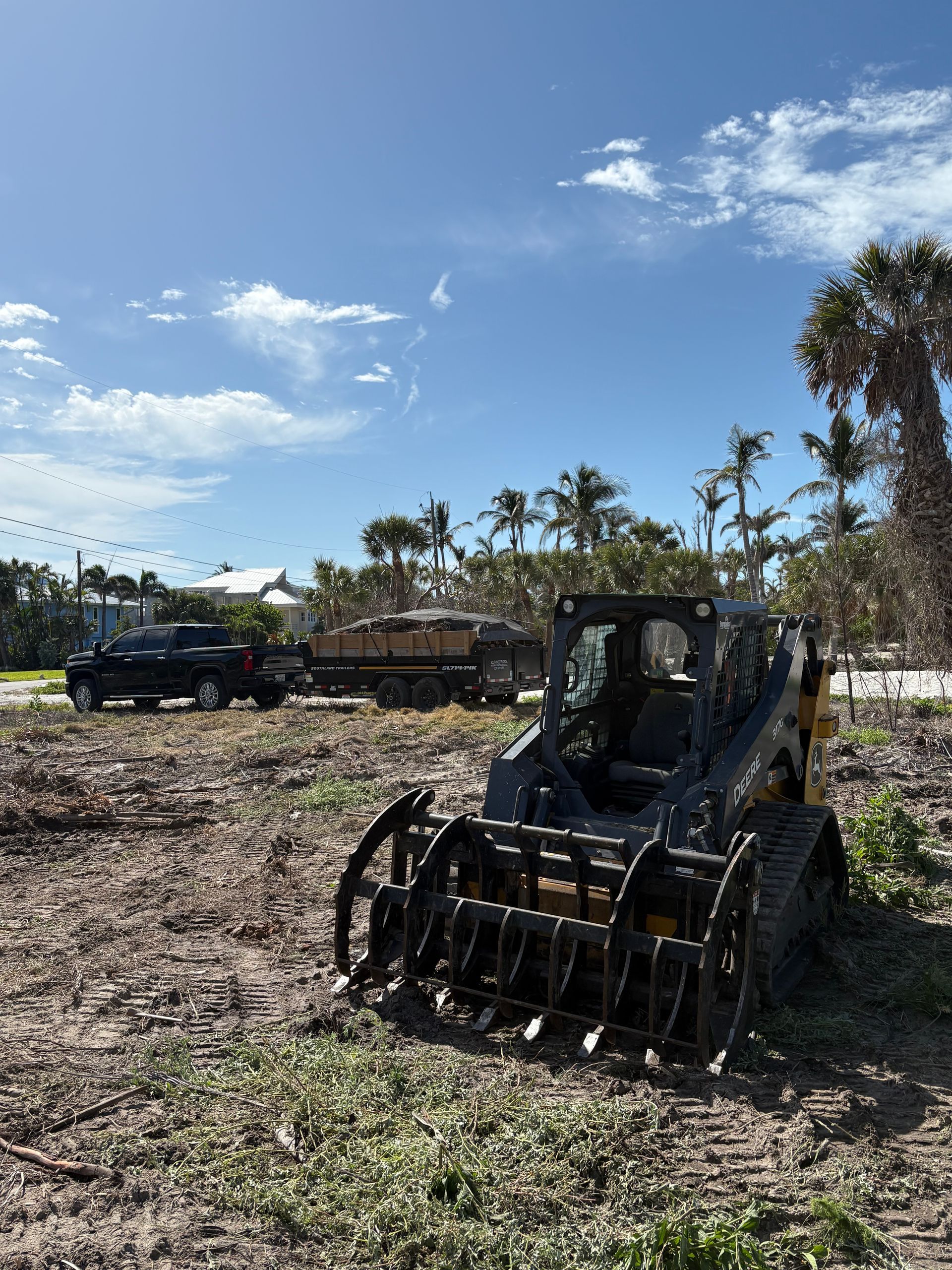 Yellow and black skid steer on dirt clearing, blue sky, palm trees, and truck in background.