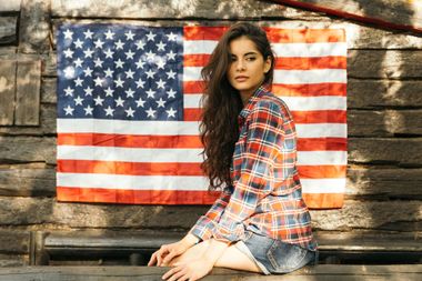 Woman sitting on a bench in front of an American flag against a rustic wooden wall