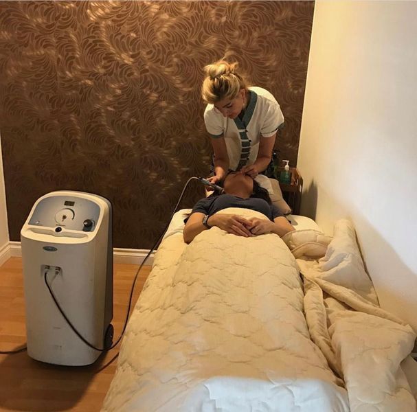 Person receiving facial treatment in a spa, with a therapist using a device. Light beige bedding and wall.