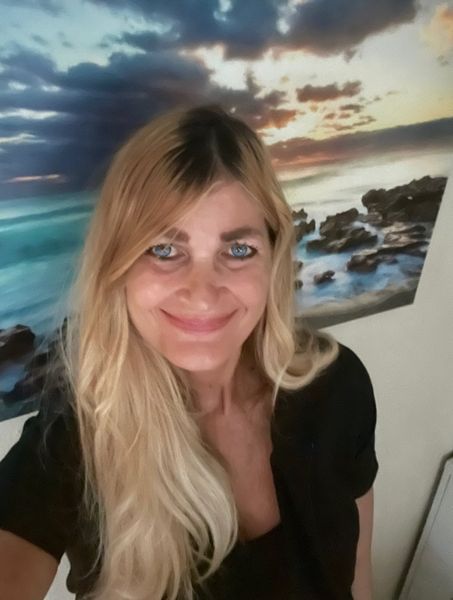 Blonde woman smiles, black shirt, against a coastal sunset painting.