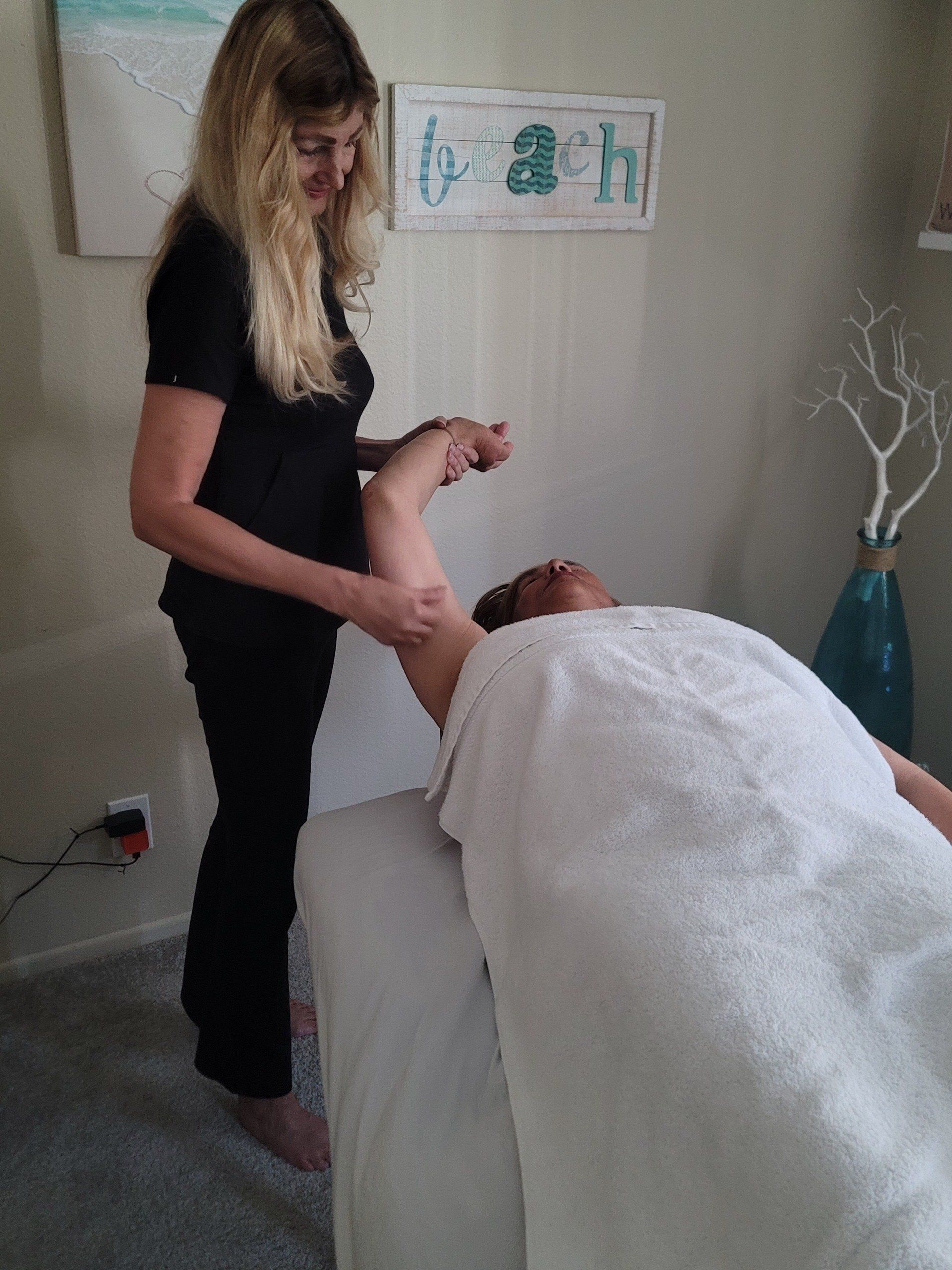 Woman giving massage to another woman's arm, indoors. Light-colored room, a massage table, and decorative items.