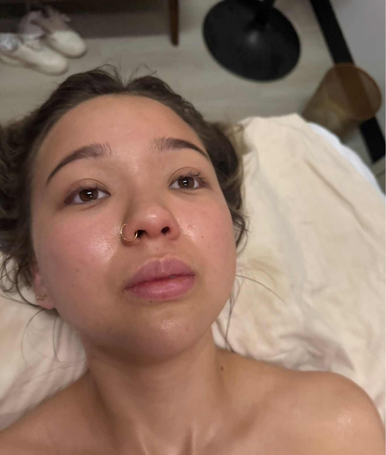 Woman with dewy skin, lying on a white surface, looking upward. A nose ring is visible.