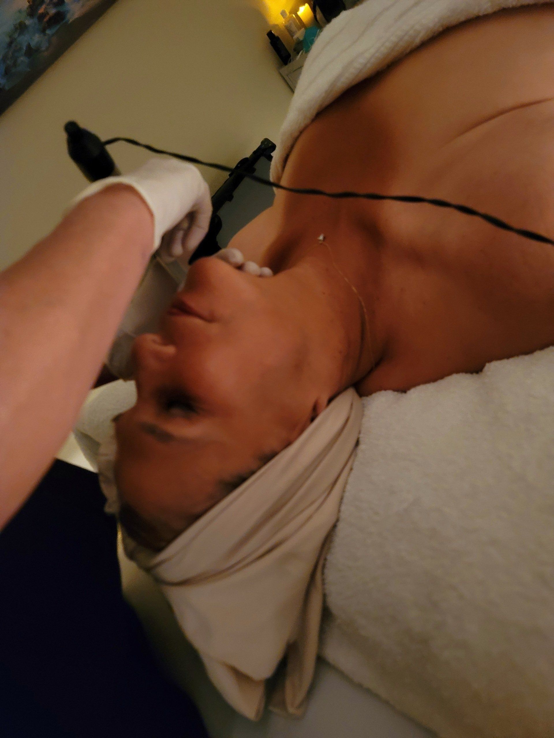 Person receiving facial treatment, wearing a towel. Gloved hand touches face. Wires present.