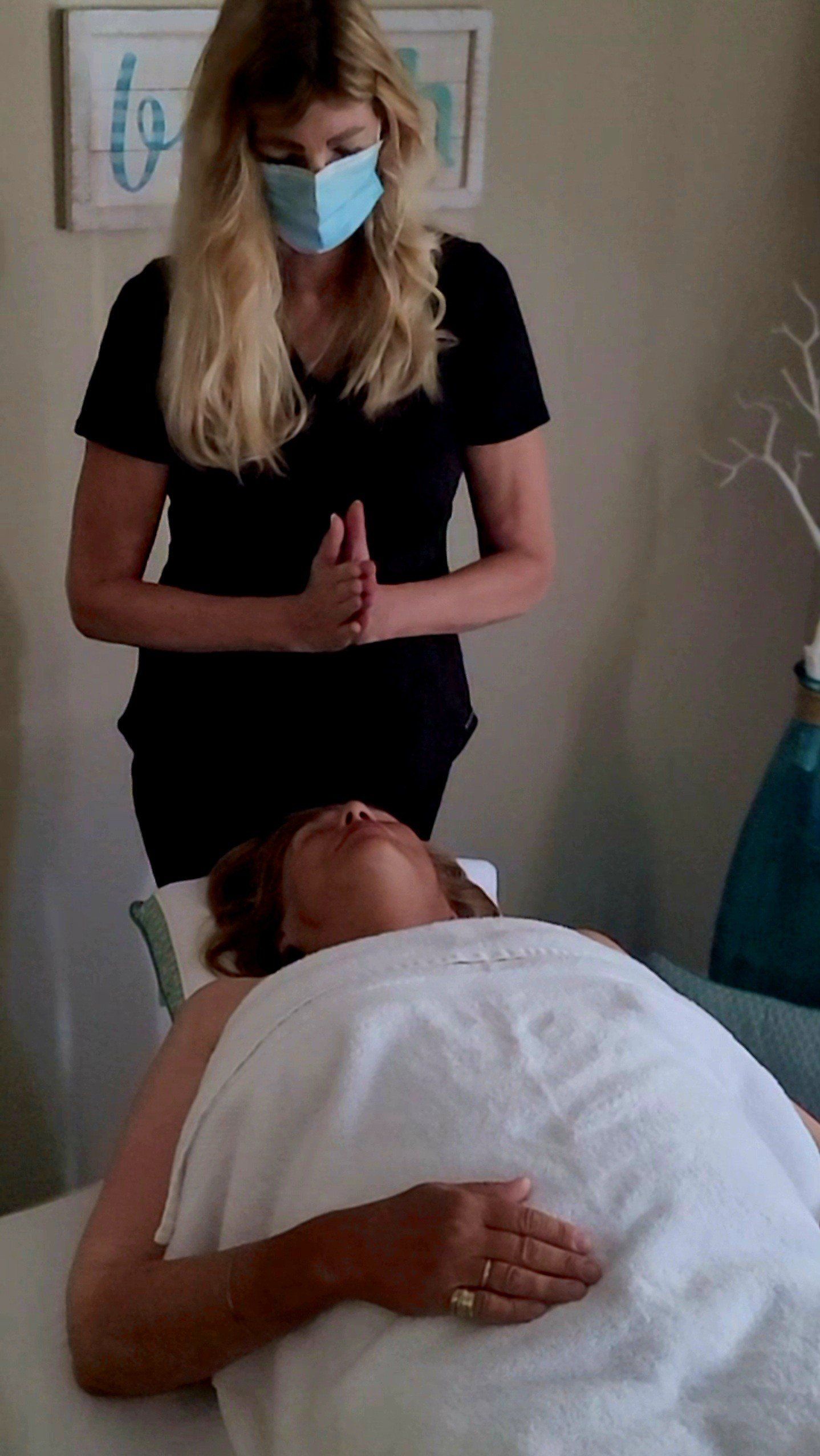 Woman in mask stands over another lying on a massage table, draped in white.