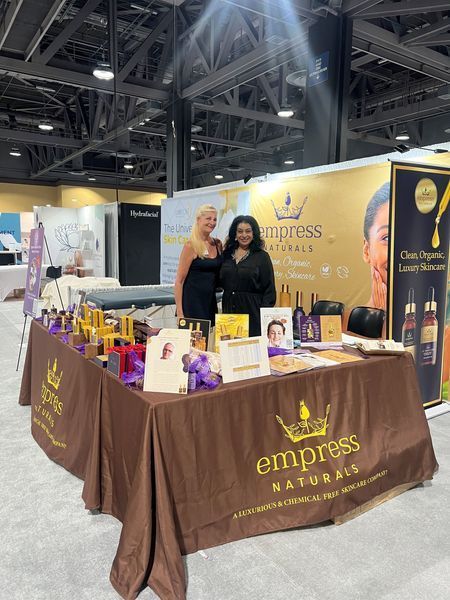 Two women stand at the Empress Naturals booth at a trade show, displaying skincare products.