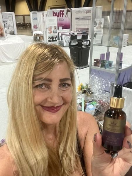 Blonde woman smiling, holding skincare product bottle at a trade show.