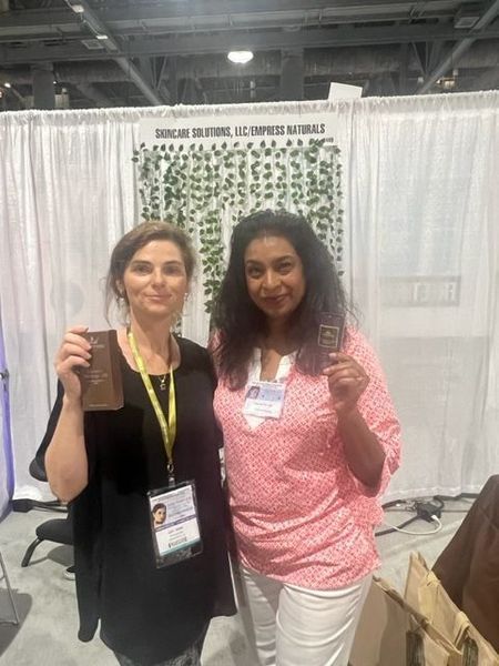 Two women holding up objects at a trade show booth, in front of a sign with greenery.
