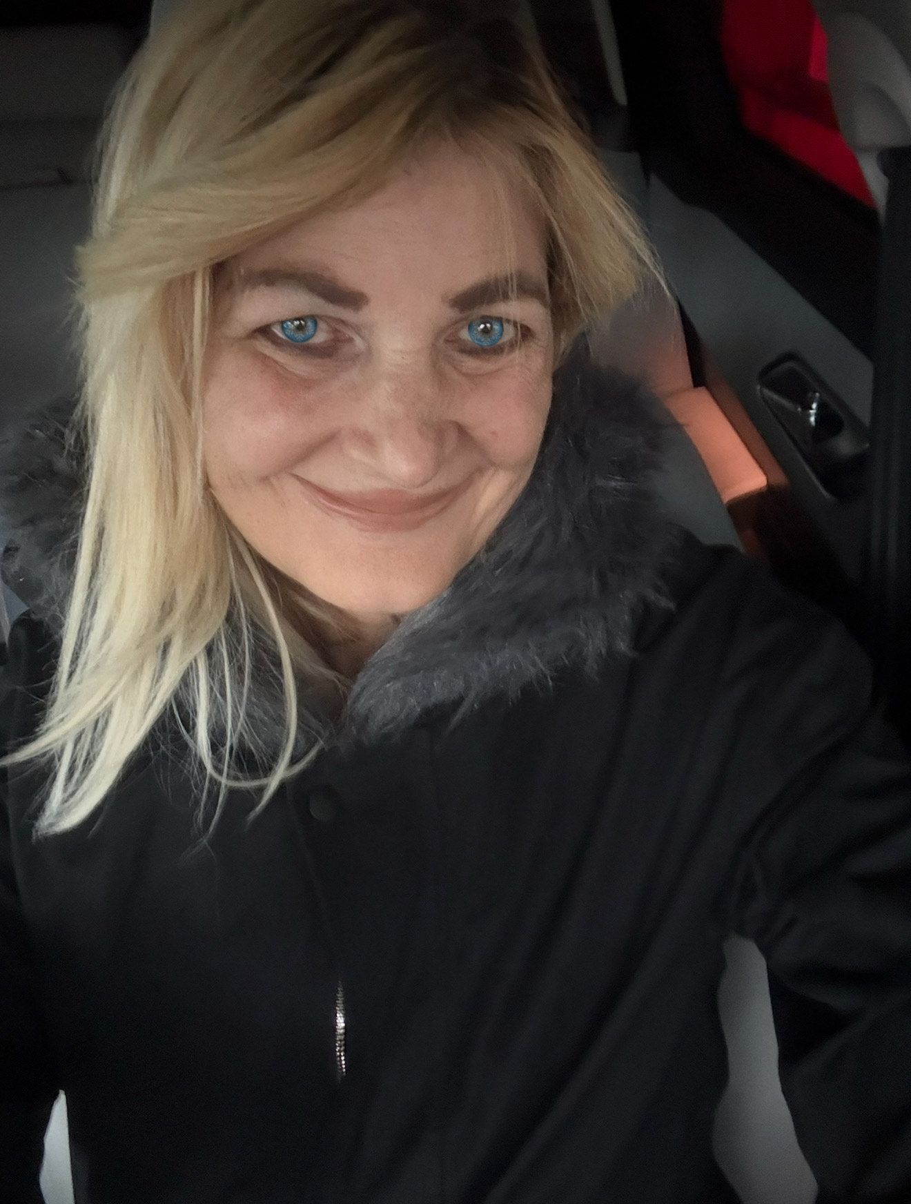 Blonde-haired person with blue eyes smiles in a car, wearing a black coat with gray fur trim.