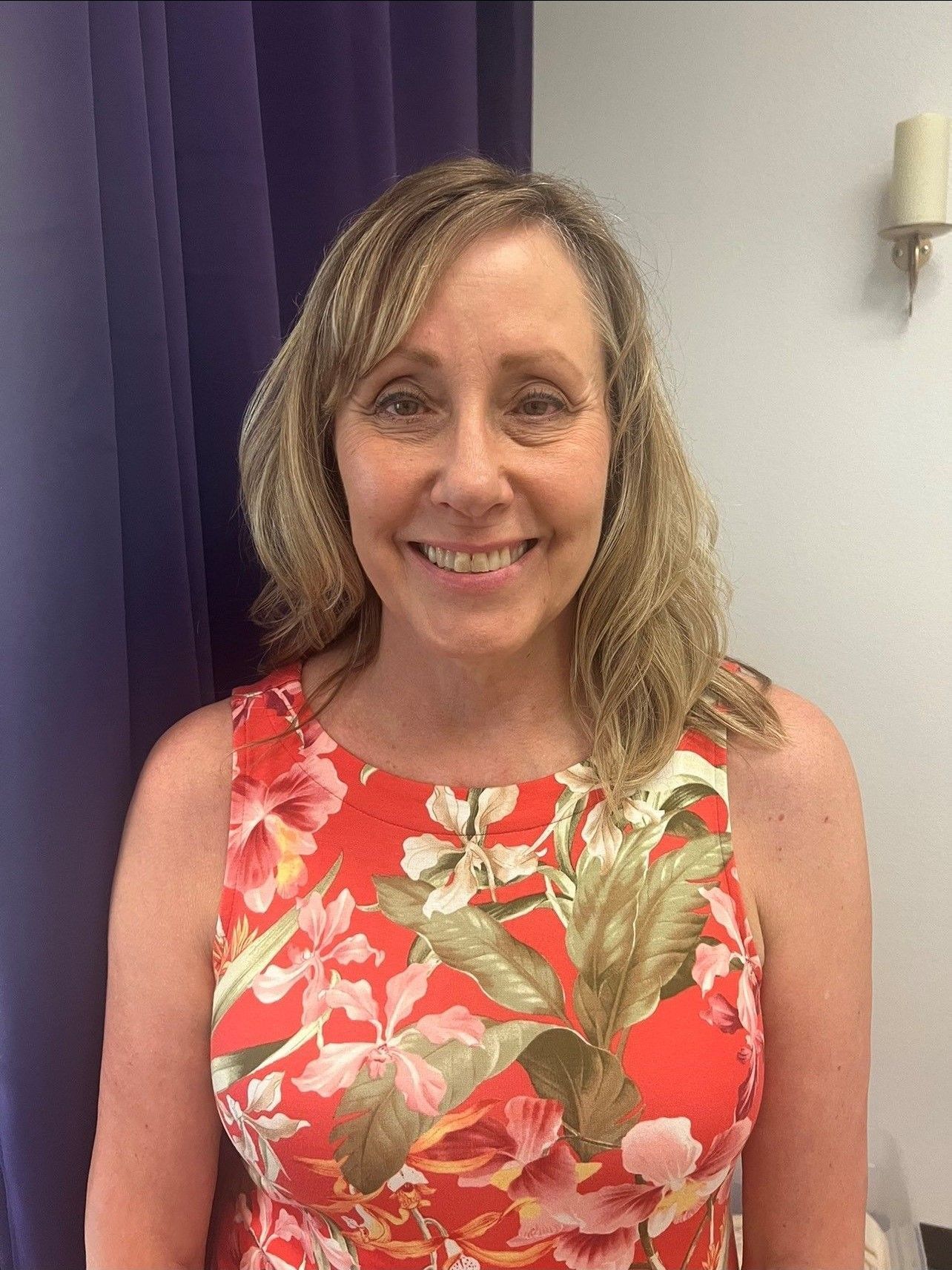 Woman in a red floral dress smiles, standing in front of a blue curtain and white wall.