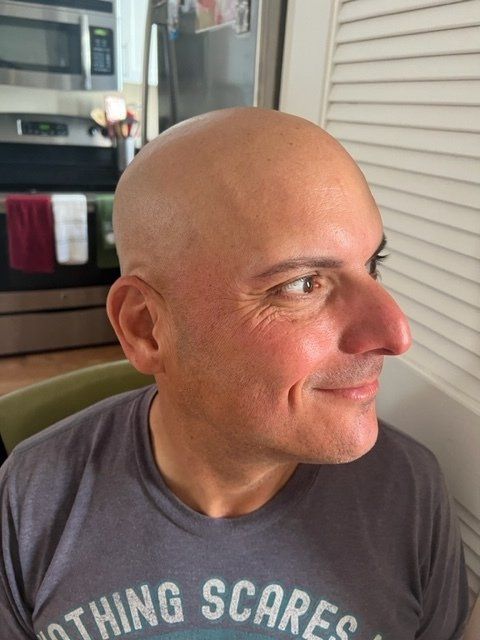 Man with a shaved head and a gray t-shirt looking right, smiling. Kitchen setting with stainless steel appliances.