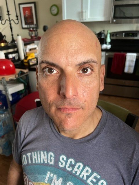 Bald man with a serious expression, wearing a gray t-shirt. Kitchen setting with appliances and decor.