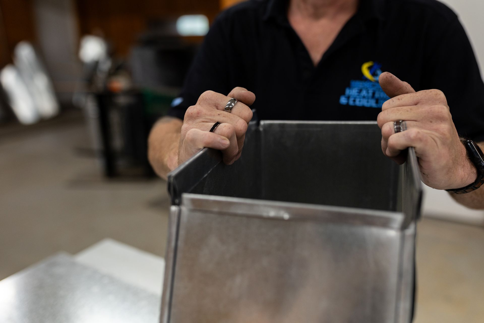 Hands holding a metal ductwork section in a workshop.
