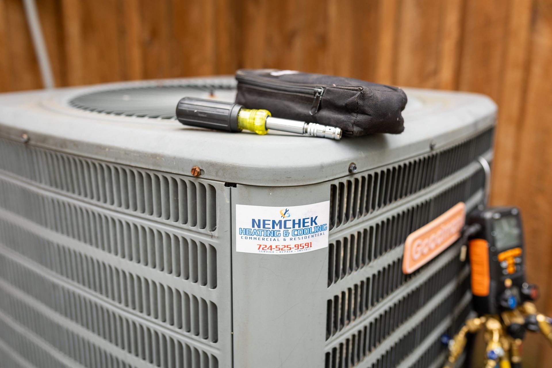 An air conditioning unit with tools on top, near a wooden fence, with a company sticker.