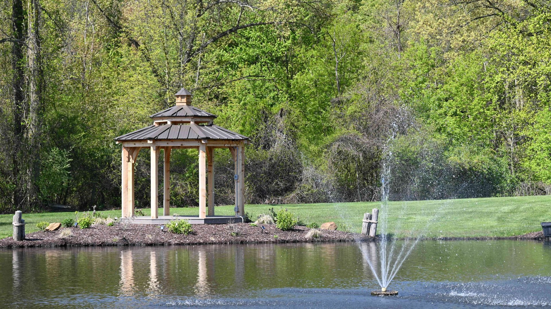 Gazebo by a pond with a fountain spraying water, surrounded by green trees and grass