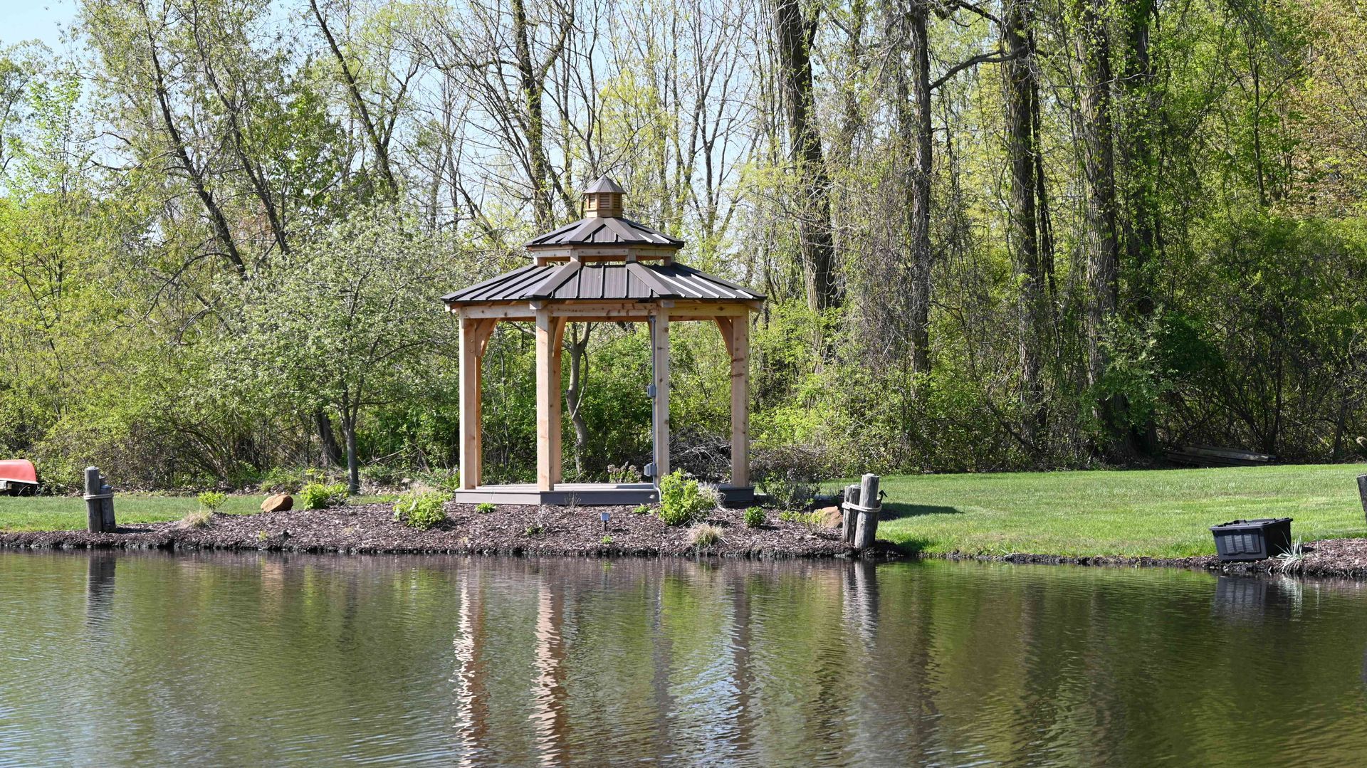 Wooden gazebo on a lakeside dock, surrounded by trees and green grass, reflected in calm water.
