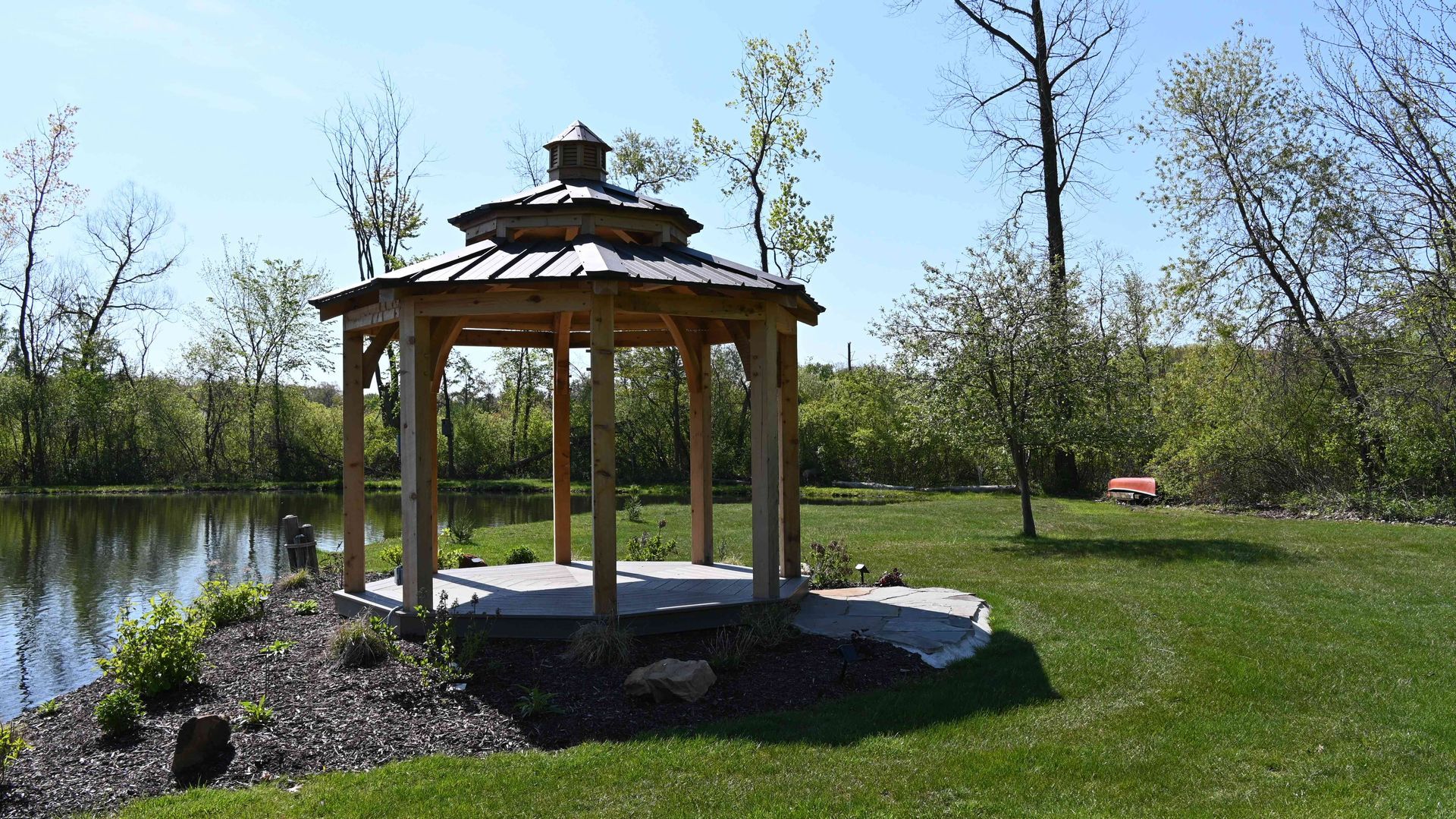 Wooden gazebo beside a pond in a sunny park with green grass and trees