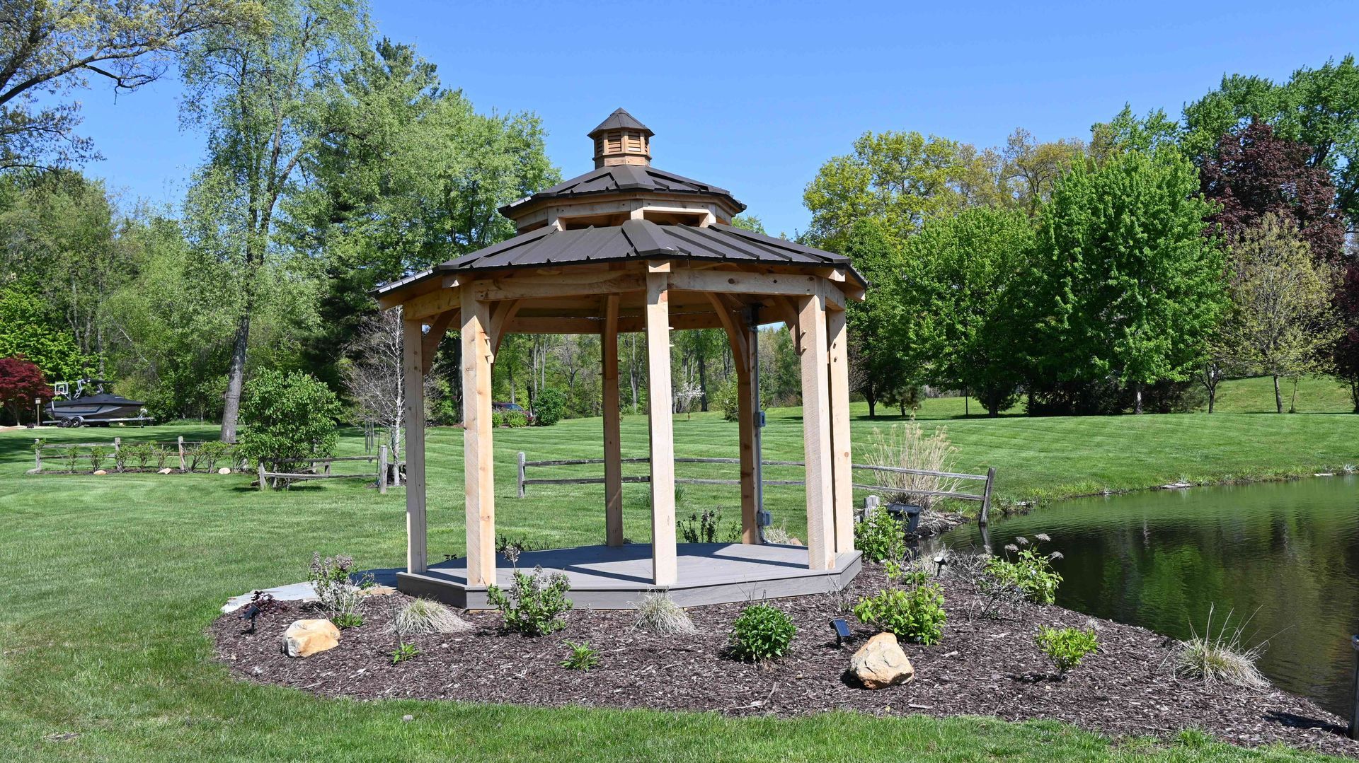 Gazebo in a landscaped park with green trees, grass, and flower beds under a clear sky