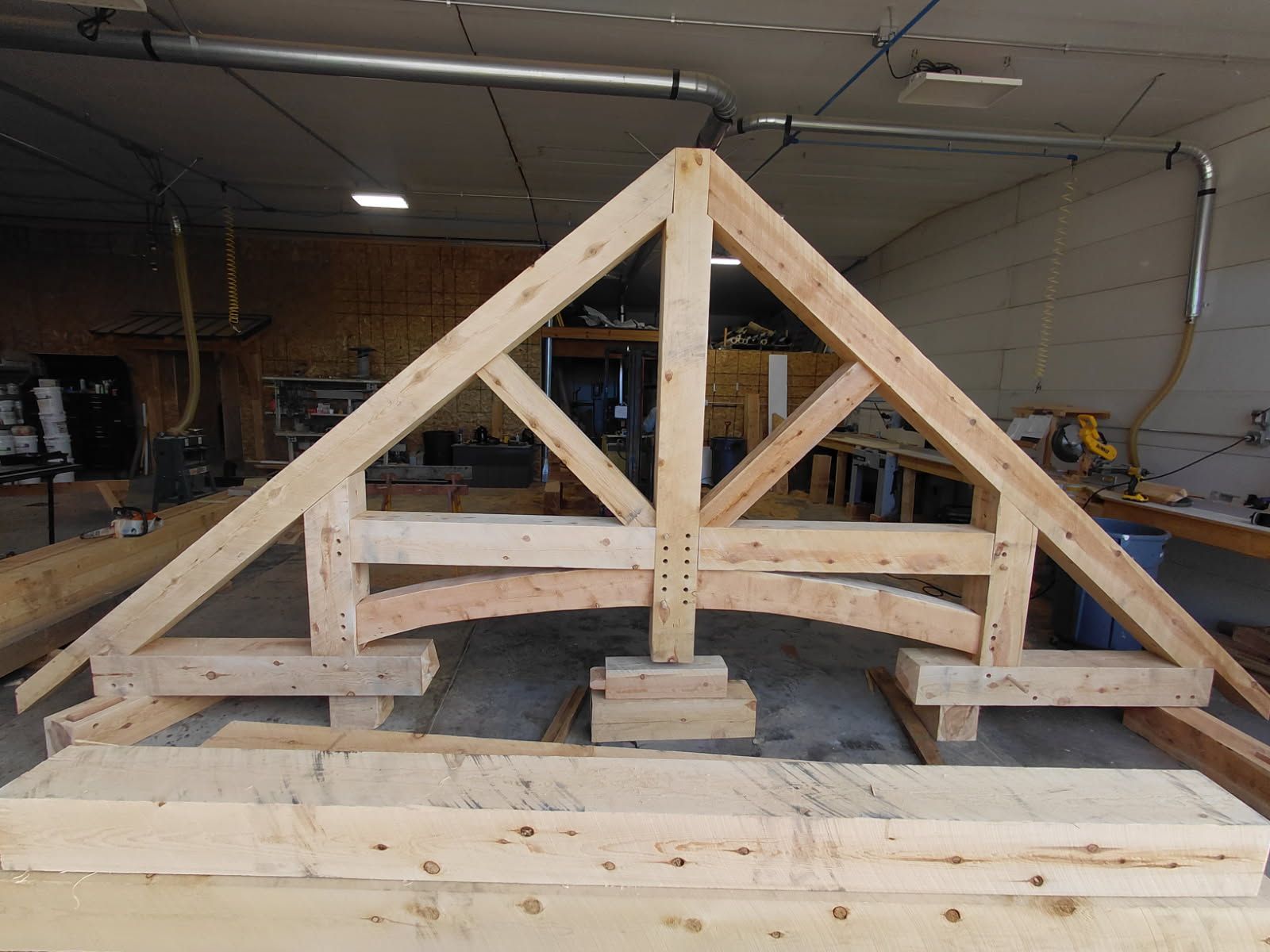 Wooden truss structure under construction in a workshop, with braces and stacked lumber on a workbench.