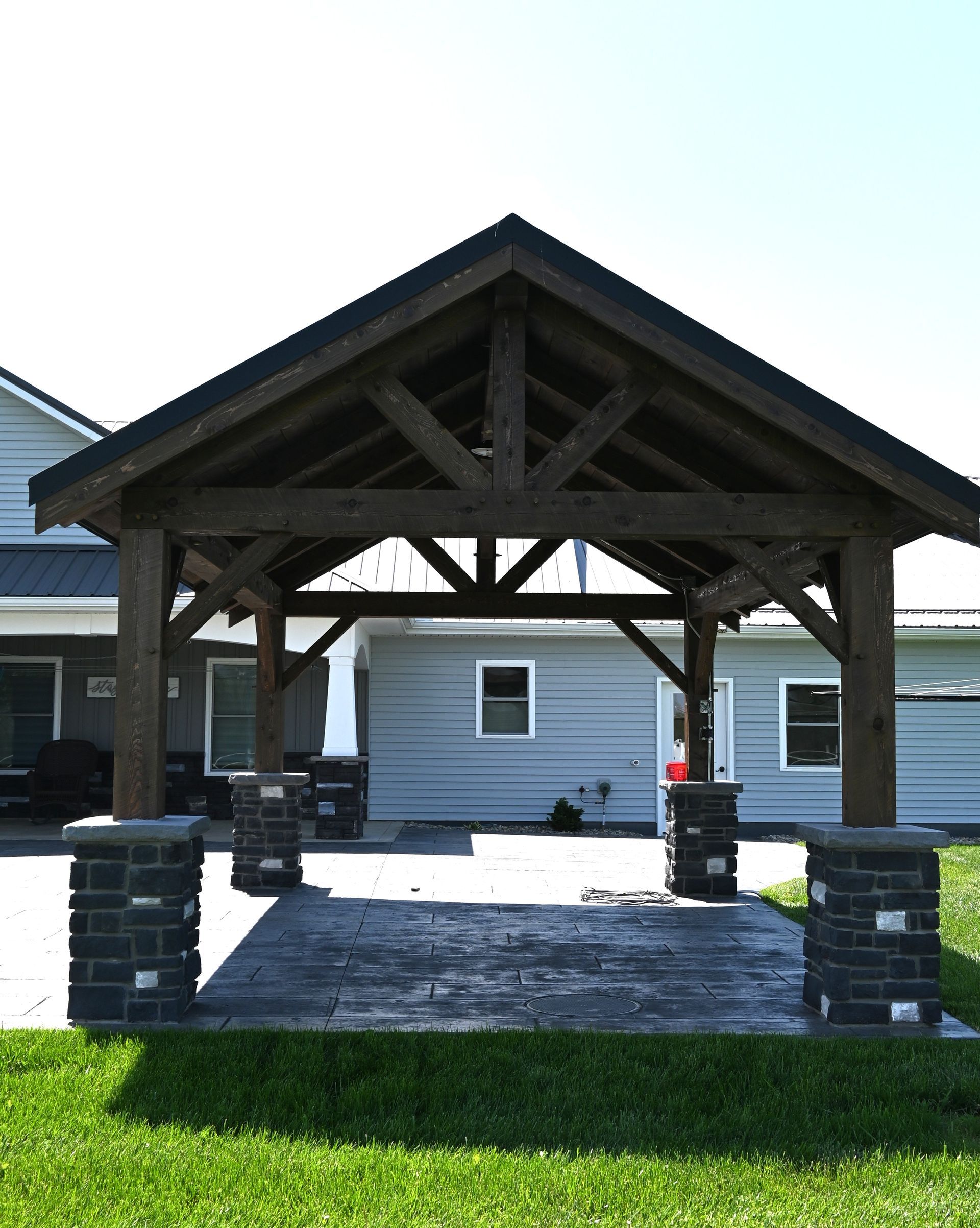 Wooden pavilion with a peaked roof over a paved walkway beside a white building and green lawn.