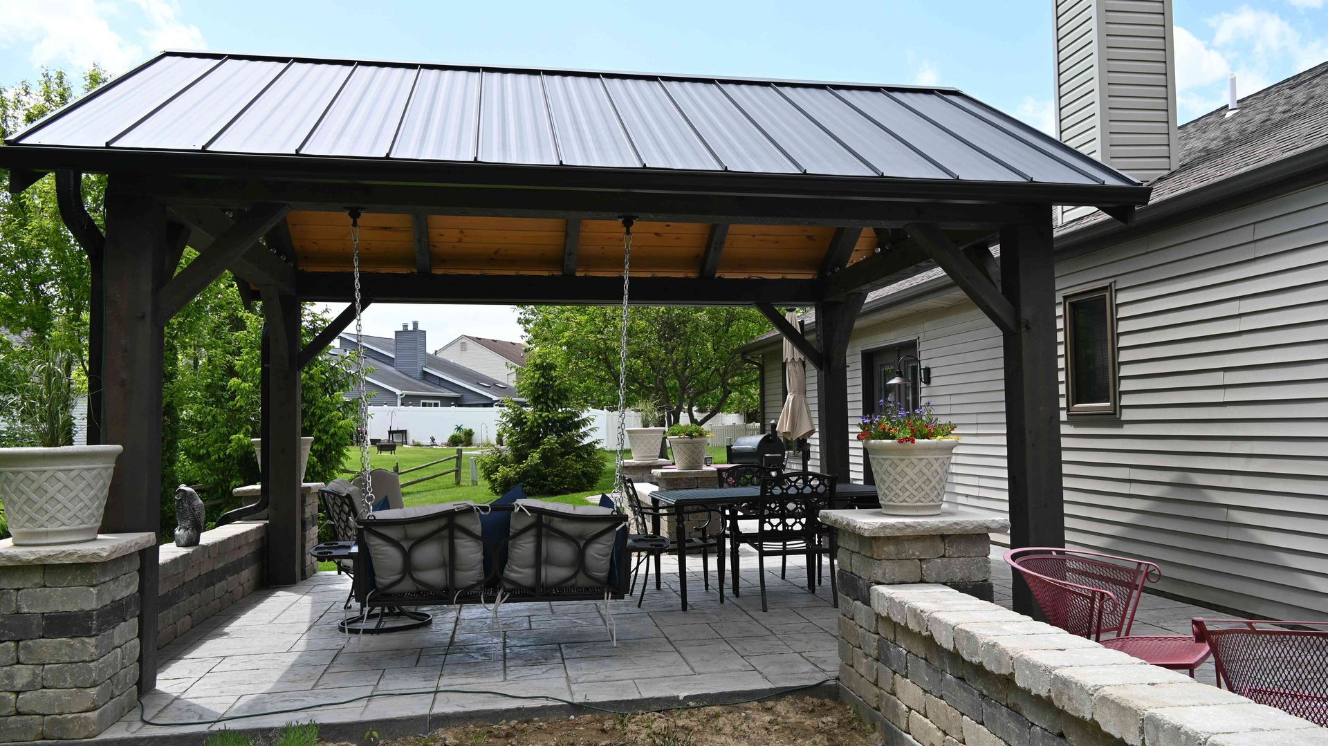Covered outdoor patio with metal roof, stone columns, and dining table beside a landscaped yard