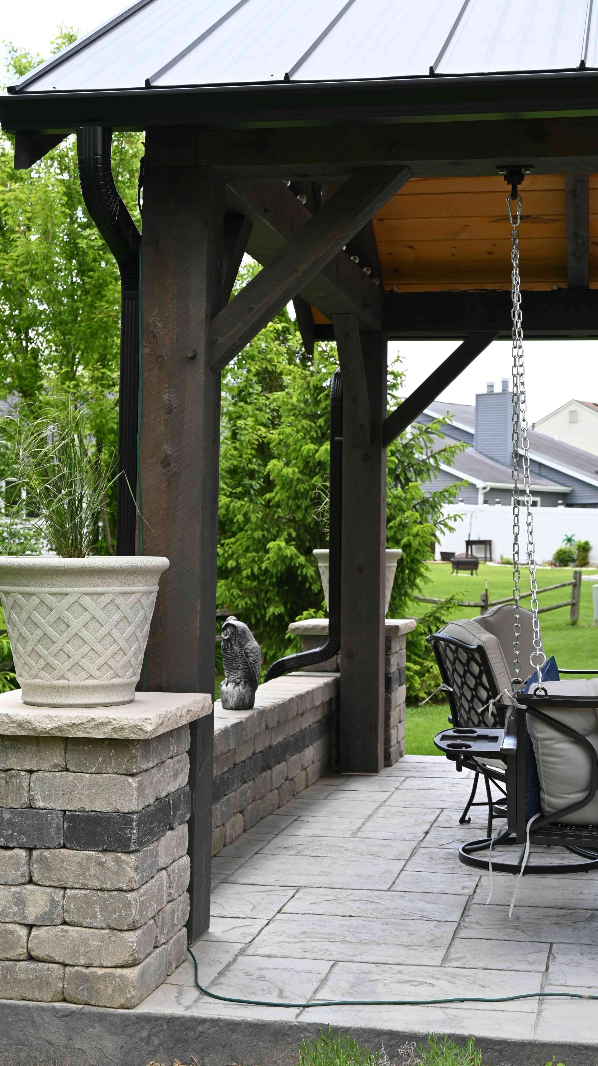 Covered stone patio with a bench, hanging swing, and potted plant beside a garden path