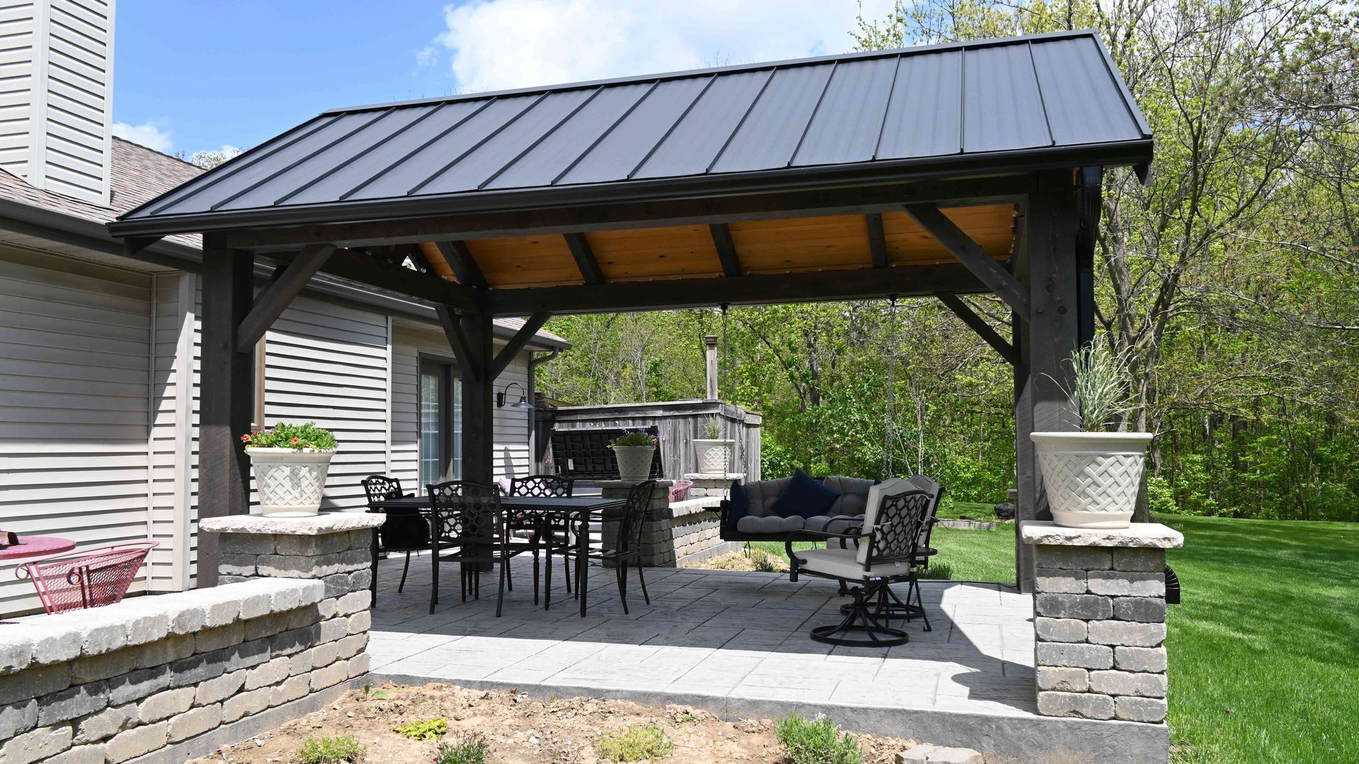 Covered backyard patio with black metal roof, outdoor dining table, chairs, and grill beside a house and lawn