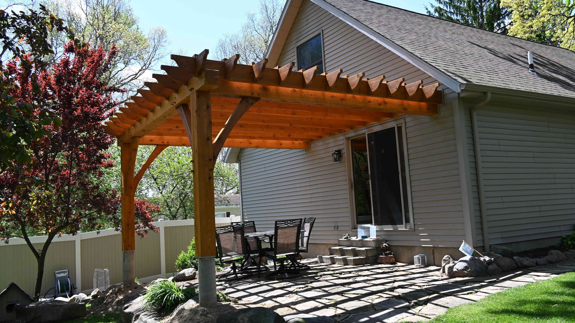 Covered wooden patio beside a house, with outdoor chairs and a table under a pergola in a sunny yard
