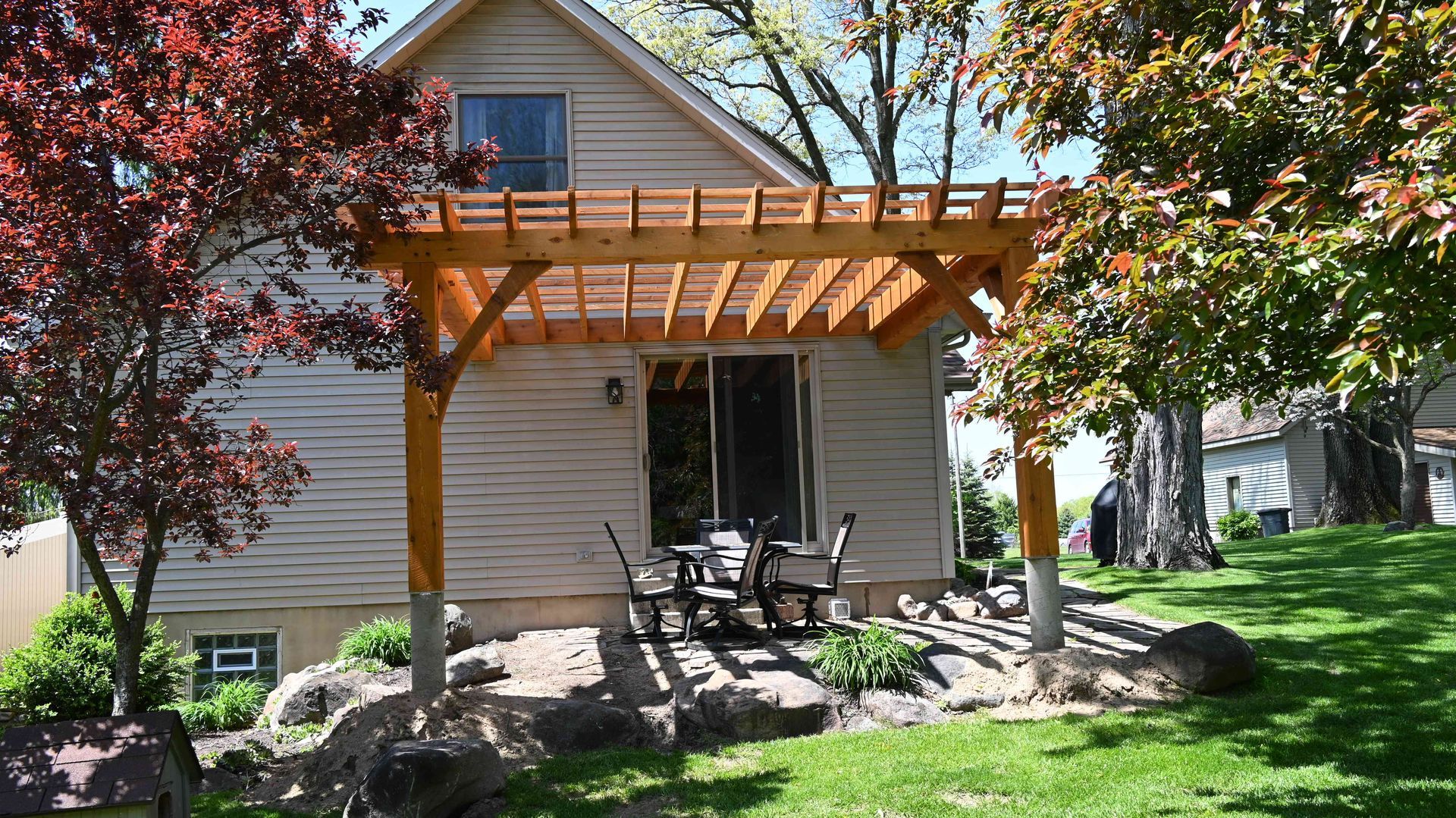 Backyard patio with wooden pergola, chairs, stone edging, and green lawn beside a beige house.