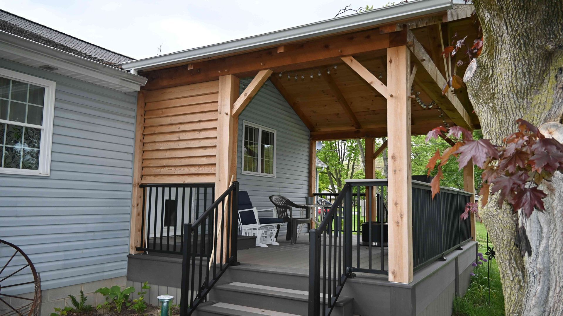 Covered front porch with wooden posts, black railings, and chairs beside a house exterior
