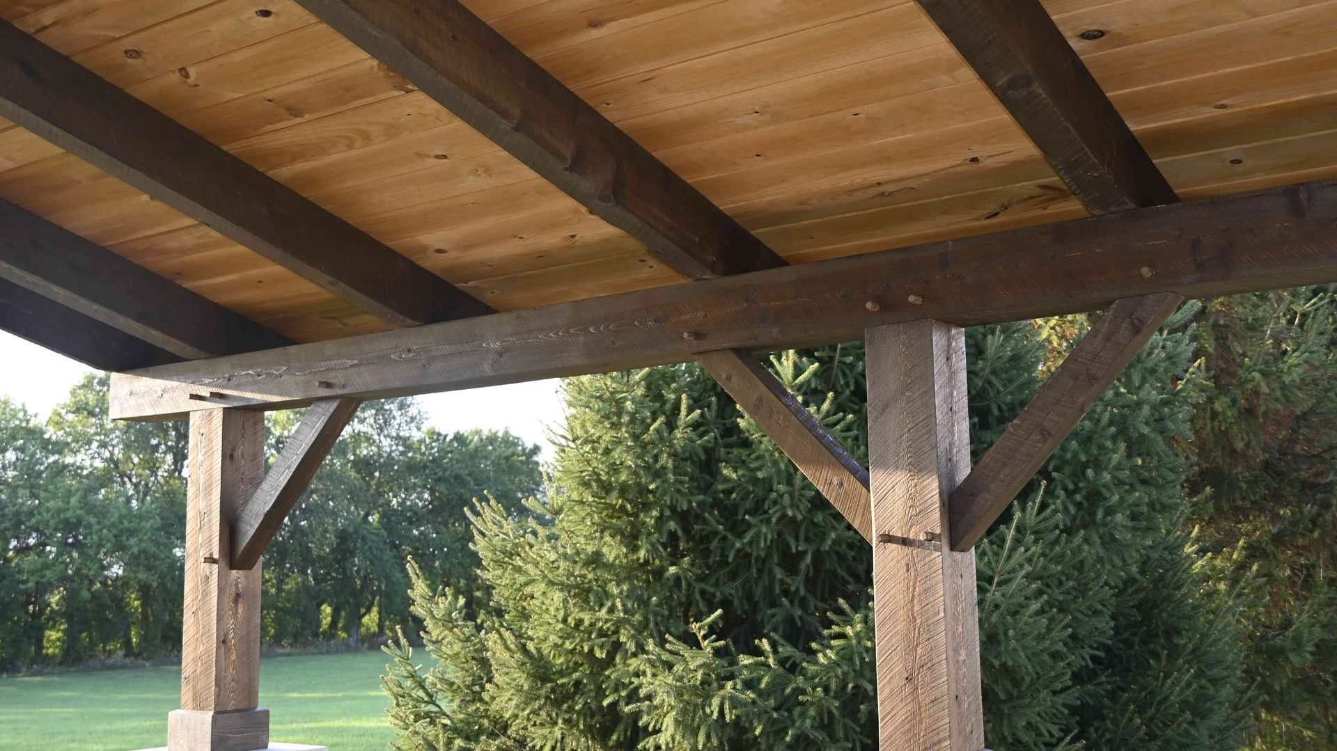 Wooden pavilion roof with thick timber posts overlooking a grassy field and trees