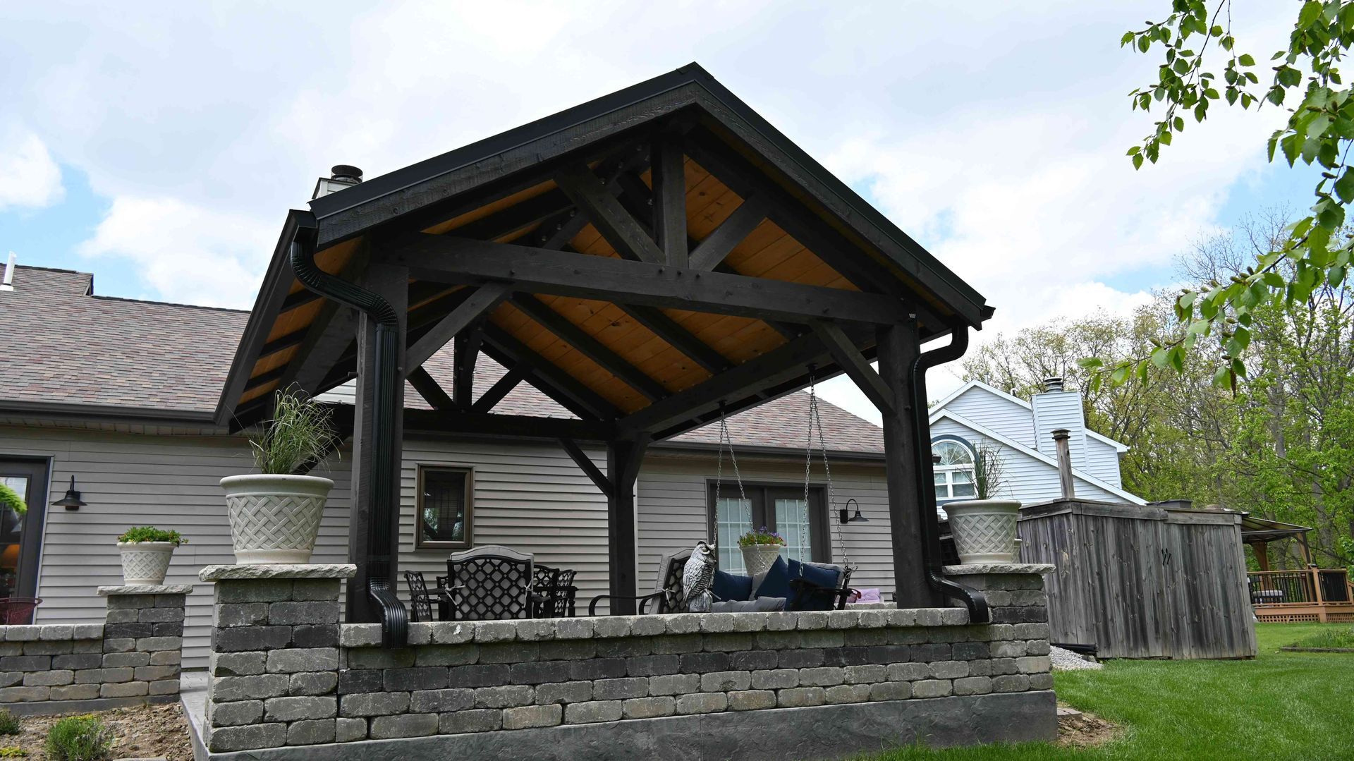 Wooden pavilion with stone base and benches in a backyard beside a house and fence