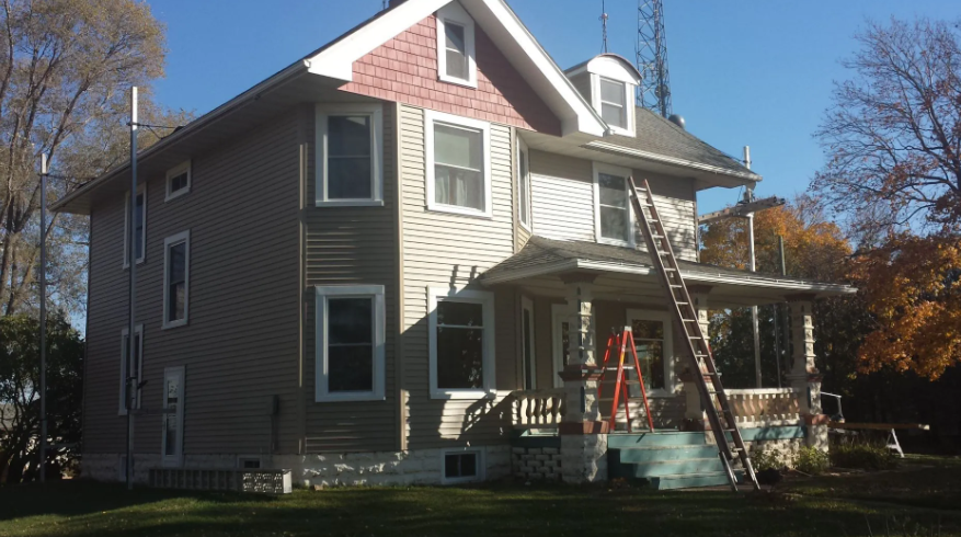 Two-story beige house with white trim and a porch, with a ladder leaning against the front in autumn.