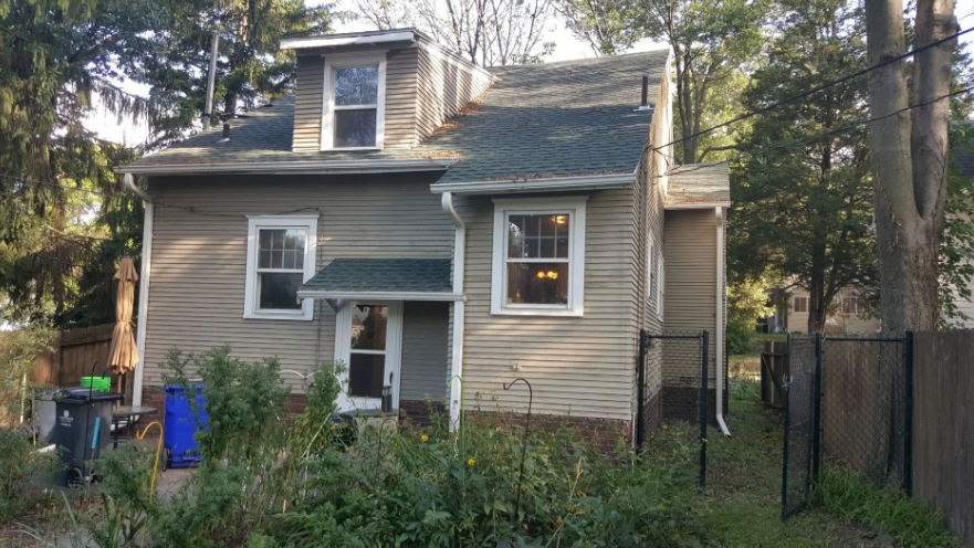 Small gray two-story house with white trim, front porch, and overgrown yard behind a black fence