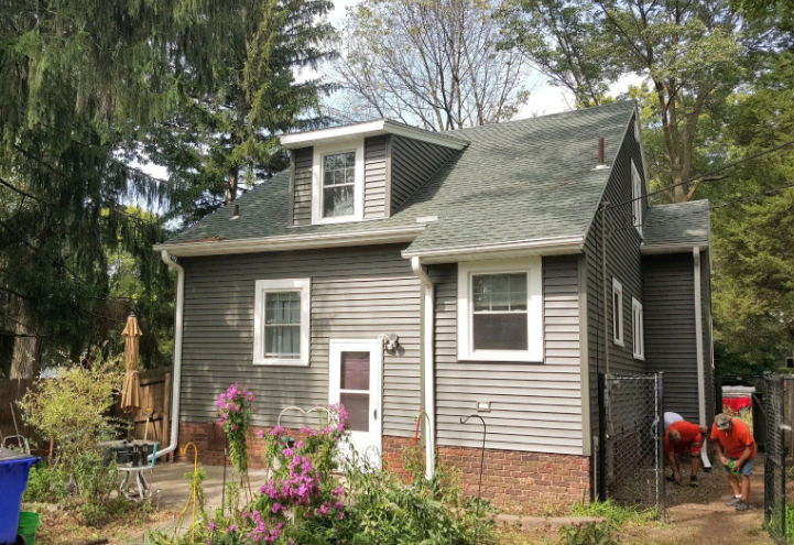 Gray house with white trim and a dormer, surrounded by trees and flowering plants