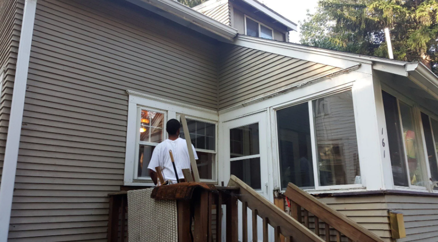 Person standing on a wooden porch facing a white-trimmed window on a gray house