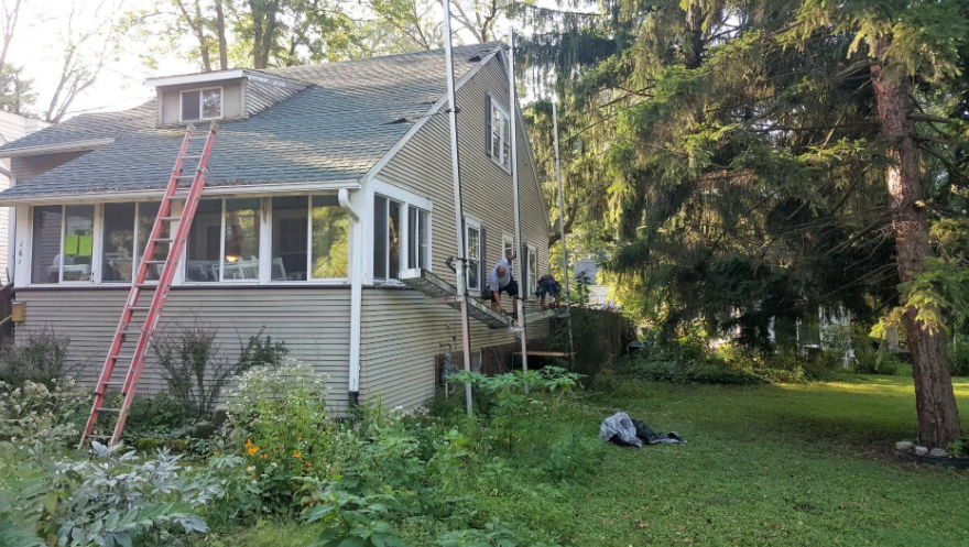 House exterior with a red ladder leaning against the roof in a grassy yard by trees