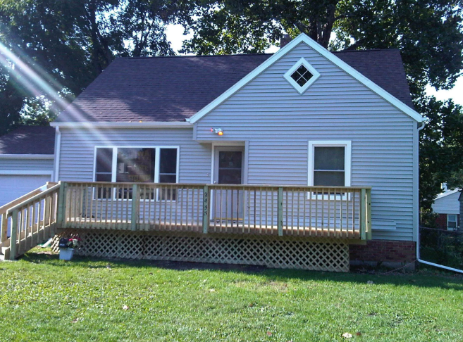 Blue house with a wooden front porch and ramp, set on a grassy lawn under trees.