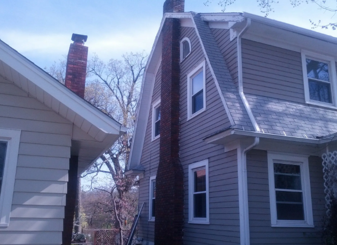 Two houses with gray siding and steep roofs, seen from a narrow side yard on a cloudy day