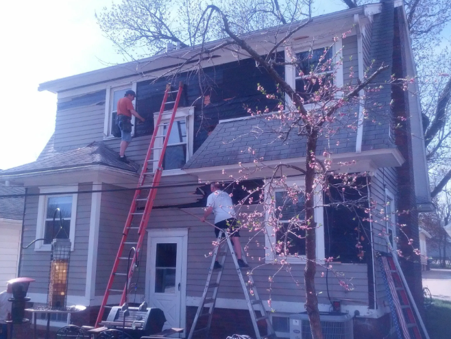 Workers on ladders trimming a large tree beside a two-story house at dusk