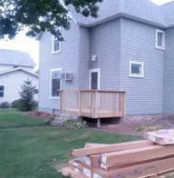Small gray house with a wooden porch deck and lumber stacks in the yard