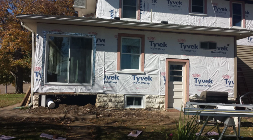 House exterior wrapped in Tyvek under renovation, with a window, door, and construction debris in front.