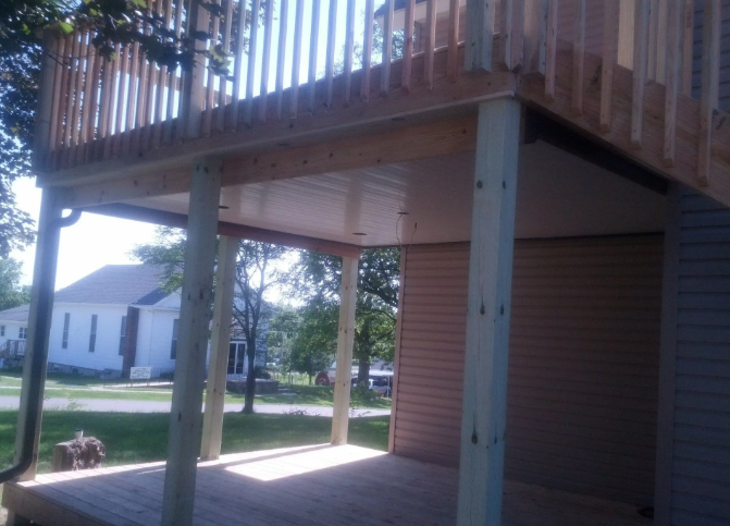Backyard under a raised wooden deck with posts, railing, and a house visible across the street.