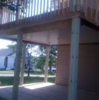 Covered outdoor walkway with wooden deck, support columns, and a view of grass and buildings beyond