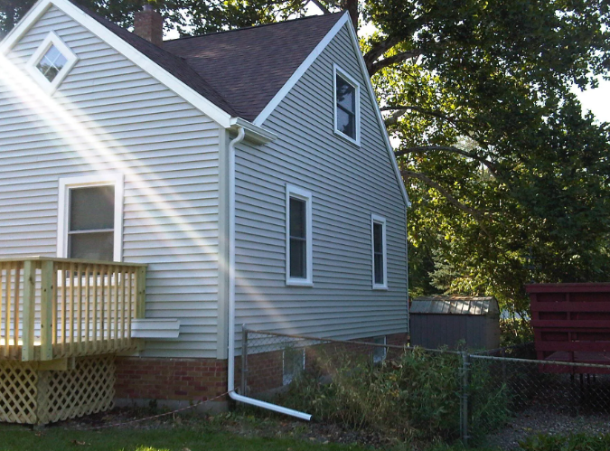 Side view of a light blue house with white trim, a small deck, and trees in the yard.