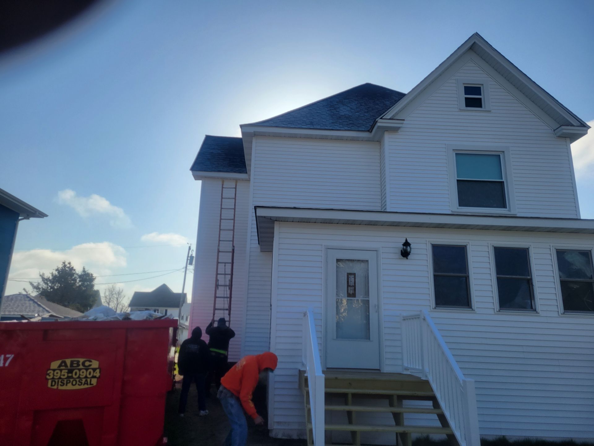 Workers beside a white house, carrying items near a red dumpster and front steps