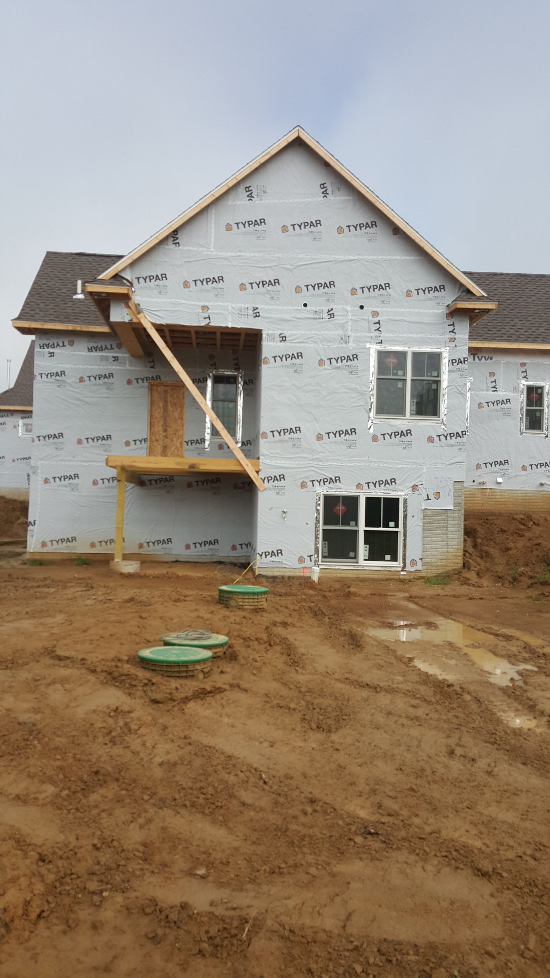 Unfinished two-story house under construction with white wrap, brown roof, and muddy front yard