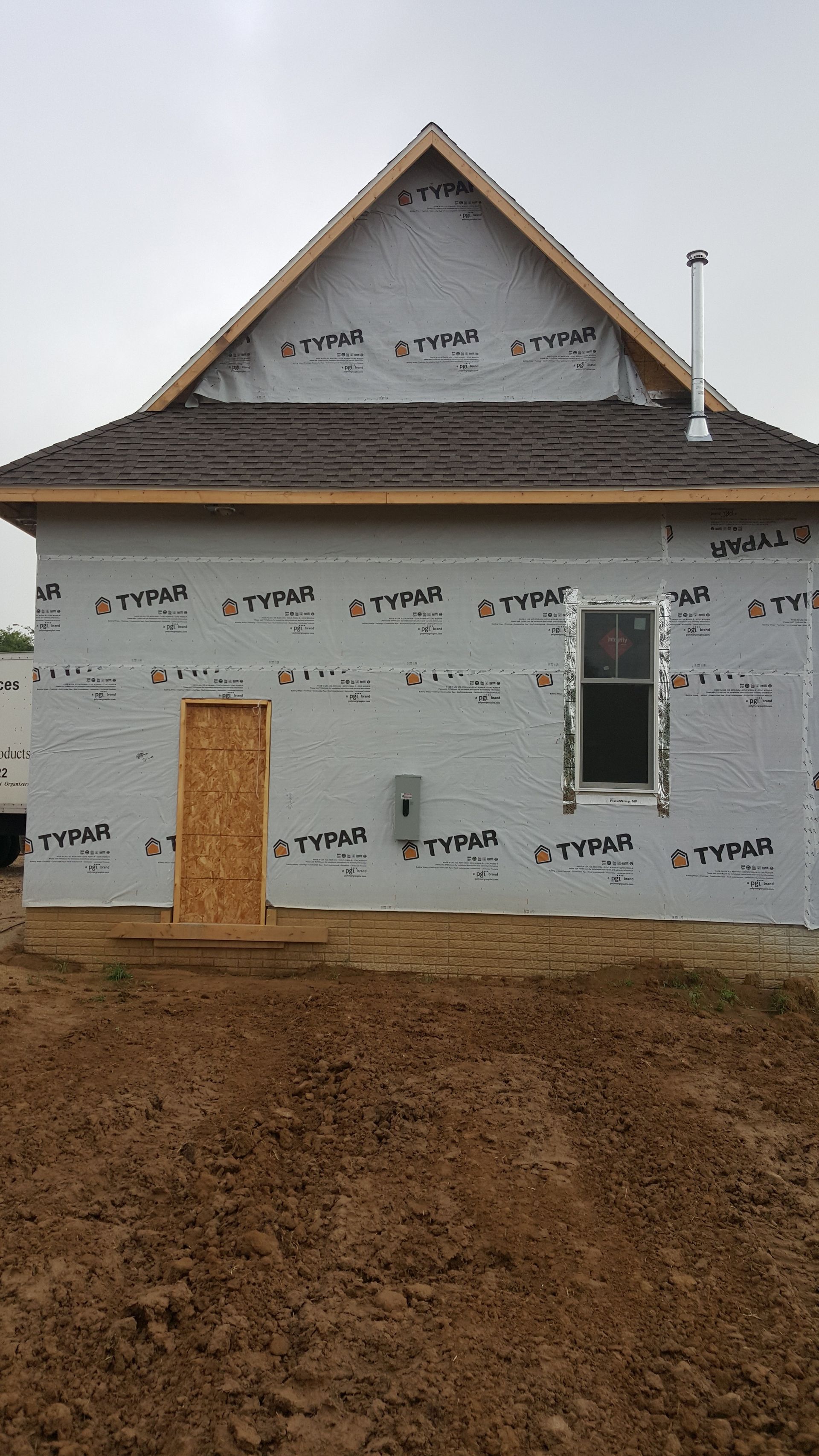 Unfinished house with white wrapped walls, brown roof, and a wooden door on a dirt lot.