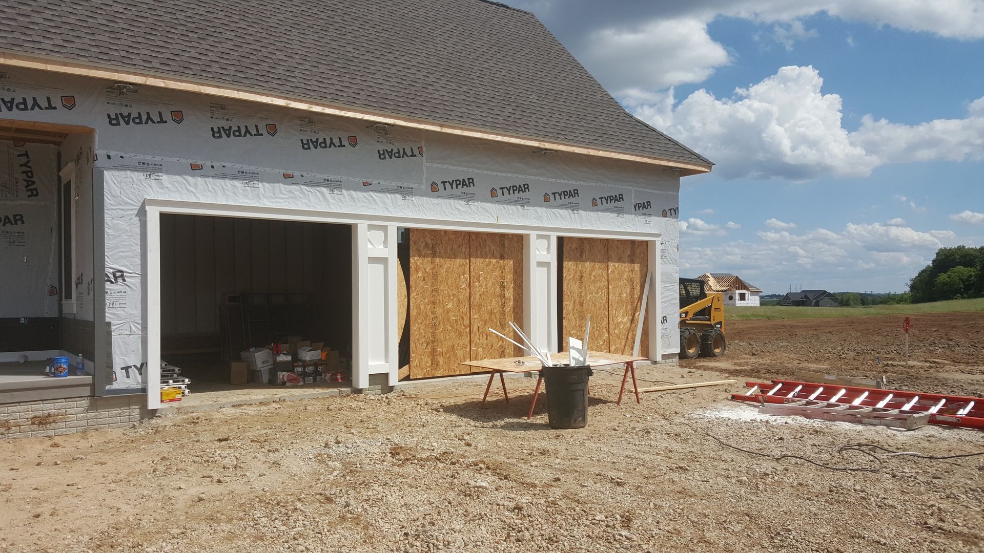 Partly built house with open garage, boarded windows, and dirt construction site under a cloudy sky